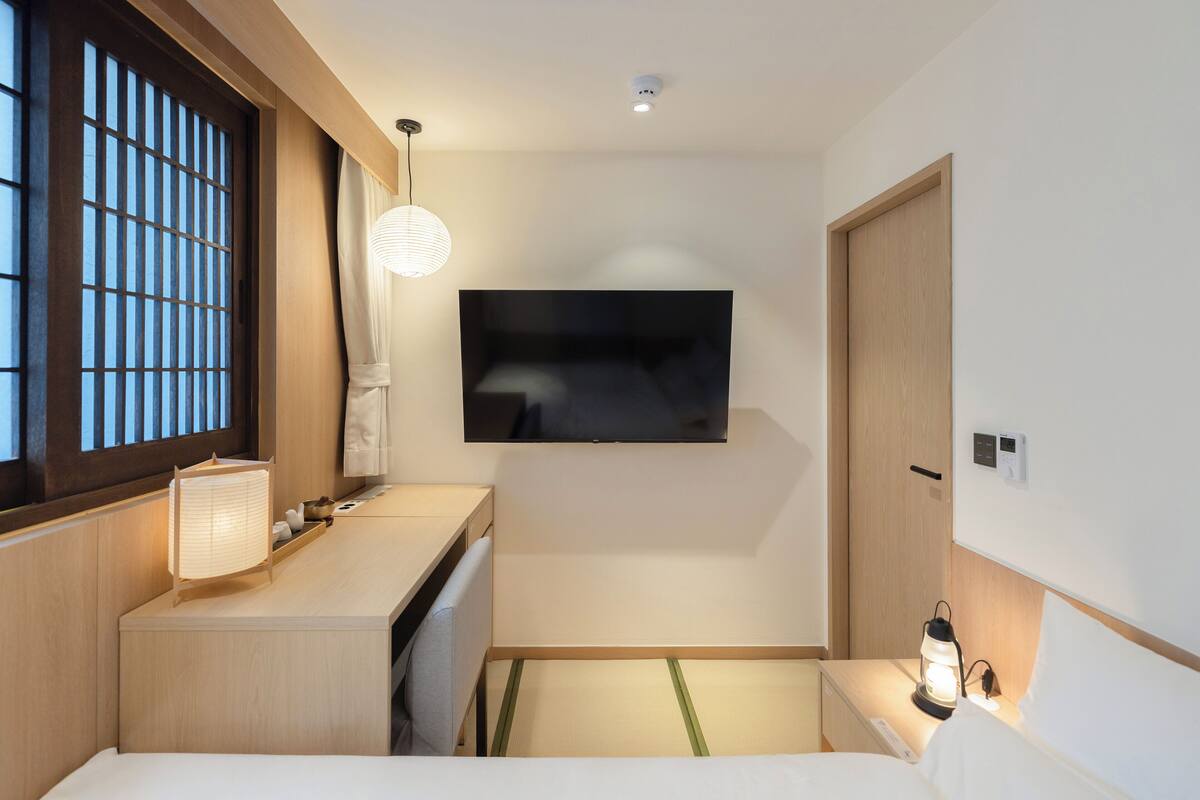 Una camera d'albergo con un letto, una TV a parete, un comodino con una lampada e una finestra vista.