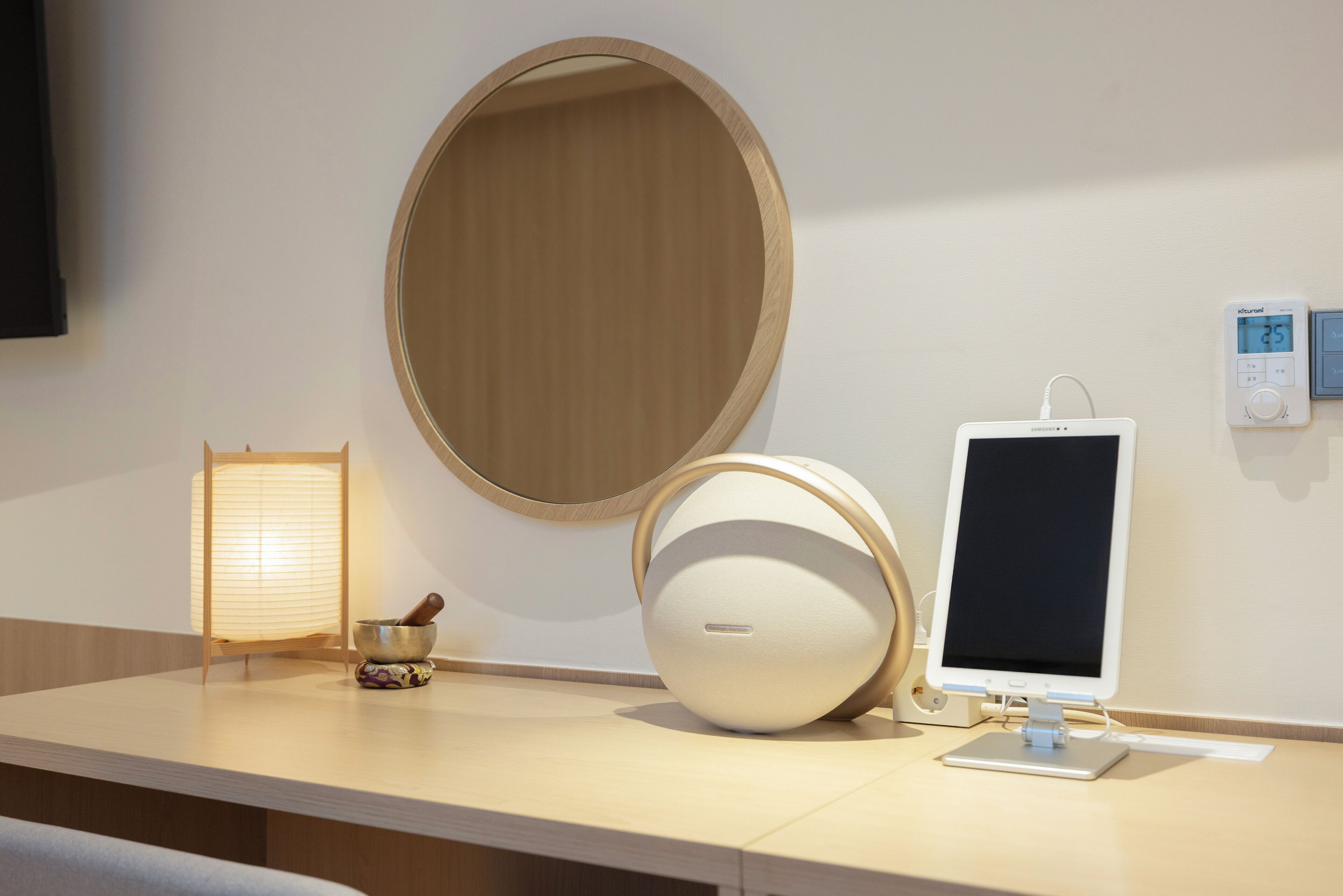 A modern room with a round mirror, a digital thermostat, and a minimalist desk setup including a lamp and a speaker.