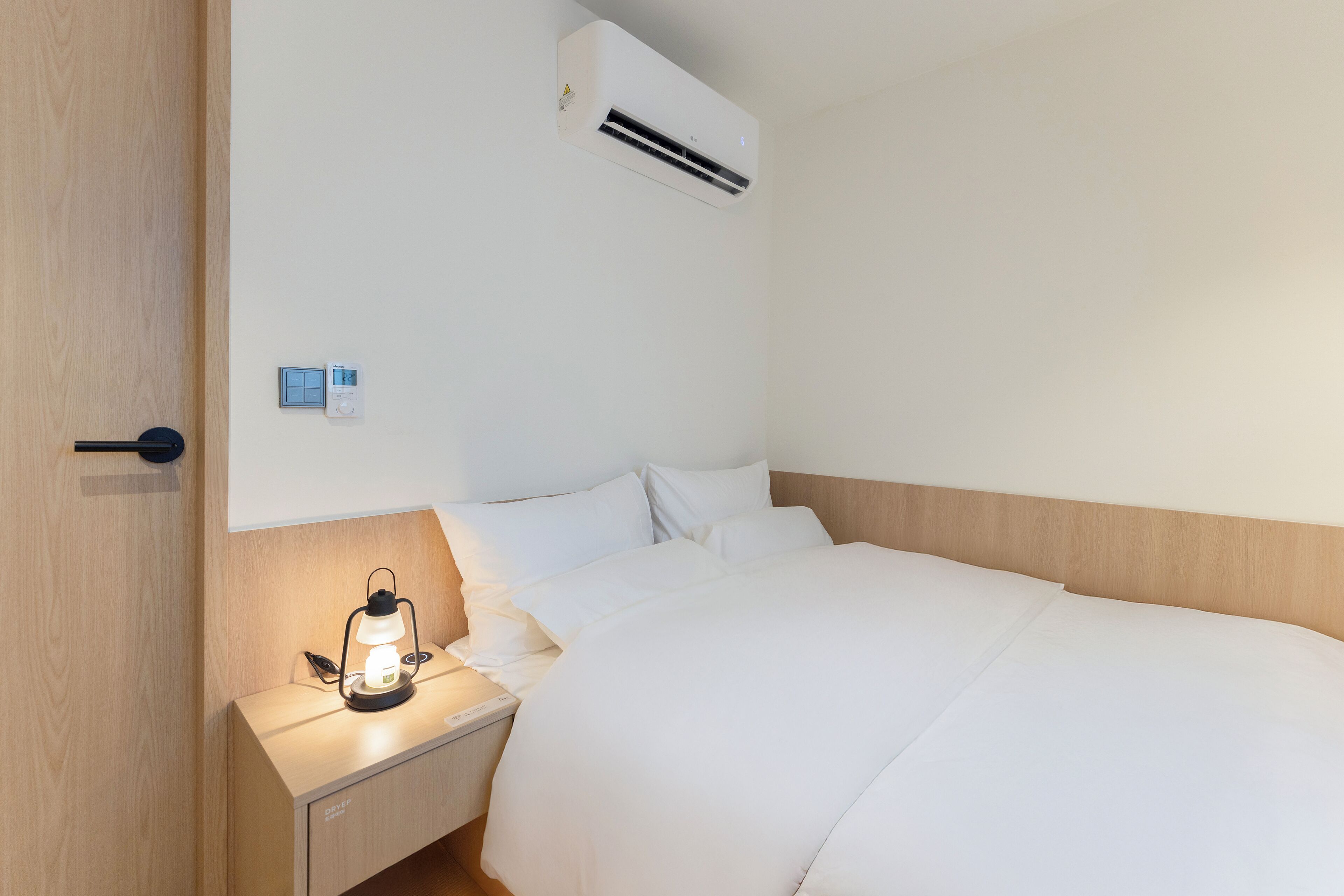 A hotel room with a bed, bedside table, lamp, air conditioning unit, and a thermostat.