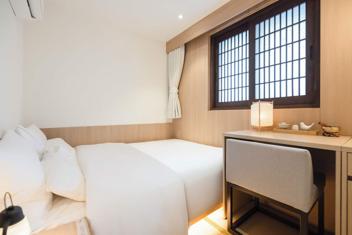 Una camera d'albergo con un letto, una scrivania, una sedia, una finestra con sbarre e una lampada da comodino.