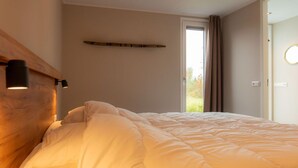 Un dormitorio con una cama grande, cabecera de madera, lámpara de noche, ventana con cortinas y puerta con interruptor de luz.