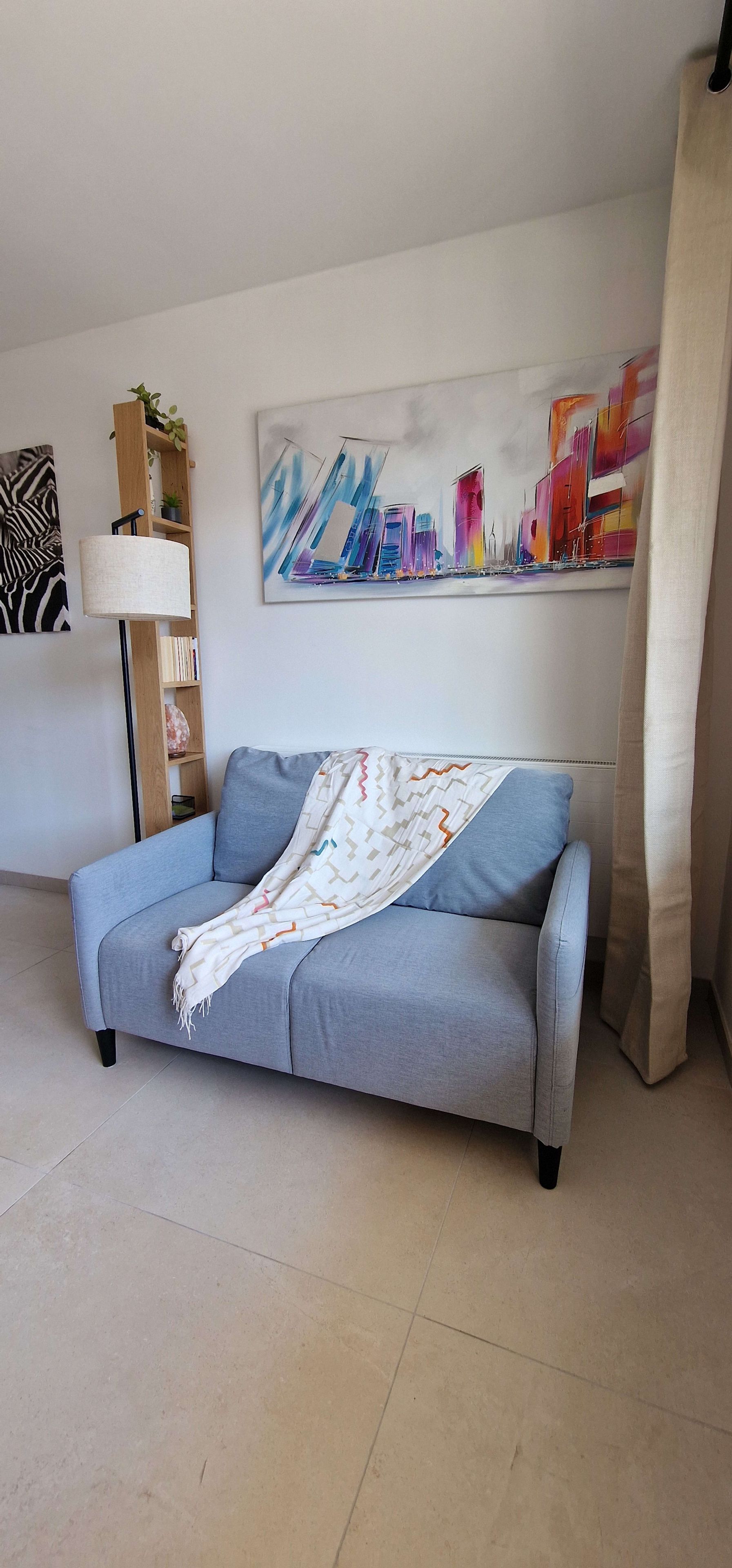 Un salon avec un canapé gris, une lampe, une bibliothèque et un tableau abstrait aux couleurs vives accroché au mur.