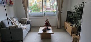 A living room with a grey sofa, a wooden coffee table with a Christmas tree, a TV on a wooden cabinet, and a window with curtains.