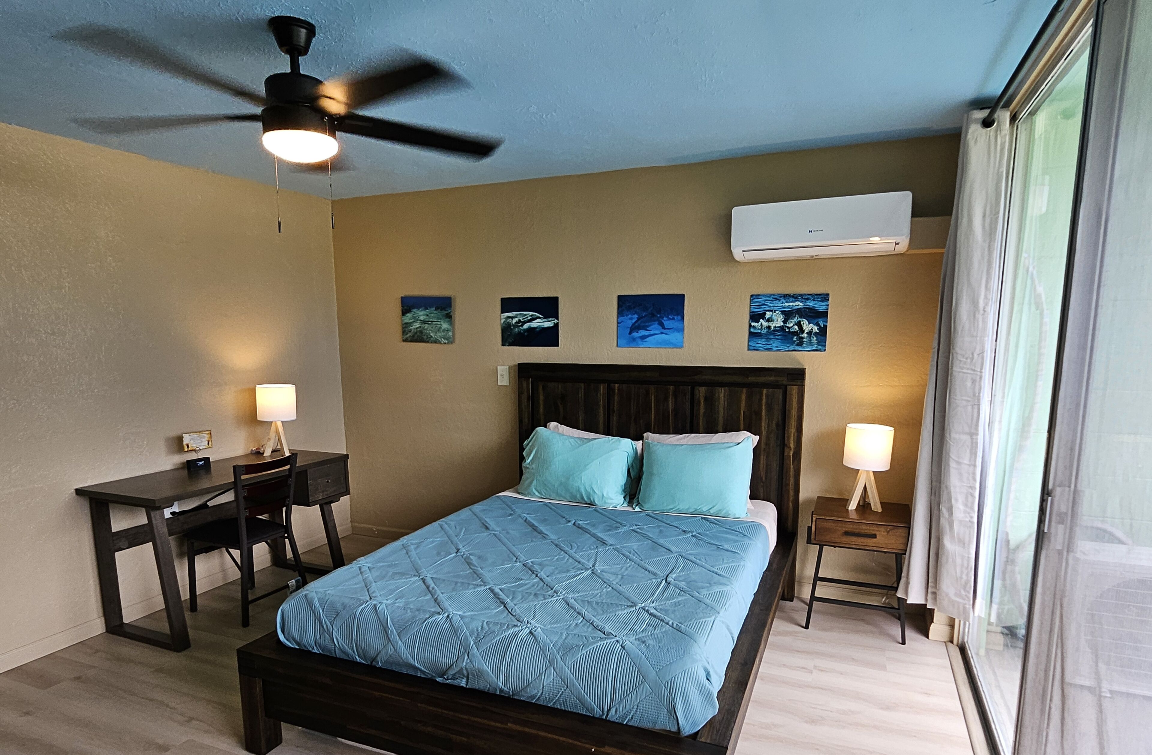 A bedroom with a bed, desk, chair, ceiling fan, and wall-mounted air conditioner.