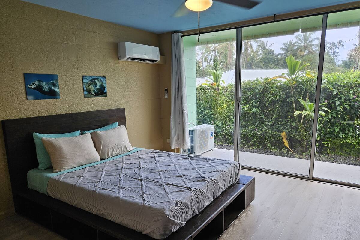 Quarto com cama grande, ventilador de teto e vista para o verde pela janela.
