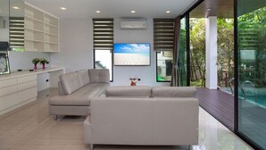 A modern living room with a beige sectional sofa, a flat-screen TV, and a large window overlooking a pool. - Lovely Pool Villa UTK B2 near Kata Beach (Karon)
