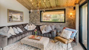 A cozy living room with a stone wall, a large window, a grey sofa, a patterned ottoman, and a decorative chandelier.
