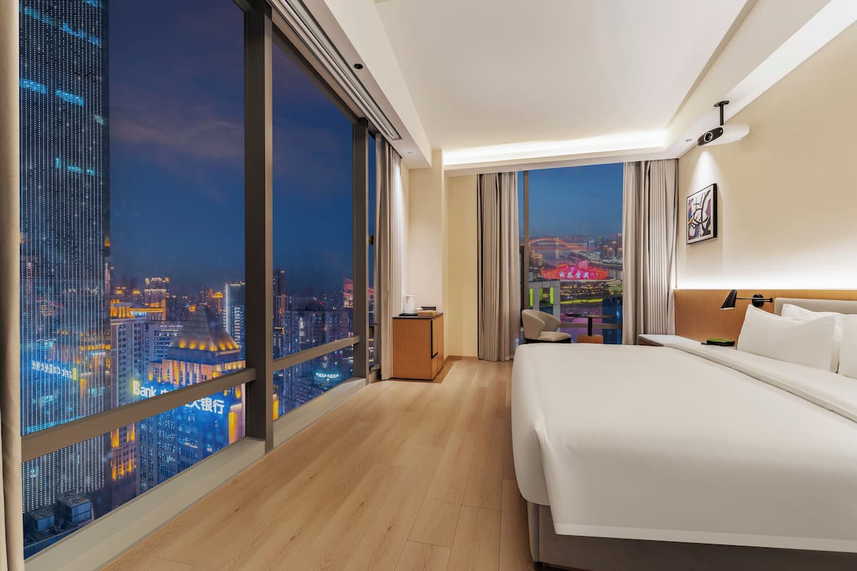 Ein Hotelzimmer mit großem Fenster und Blick auf die Stadt, einem Bett, einem Nachttisch und einem Sessel.