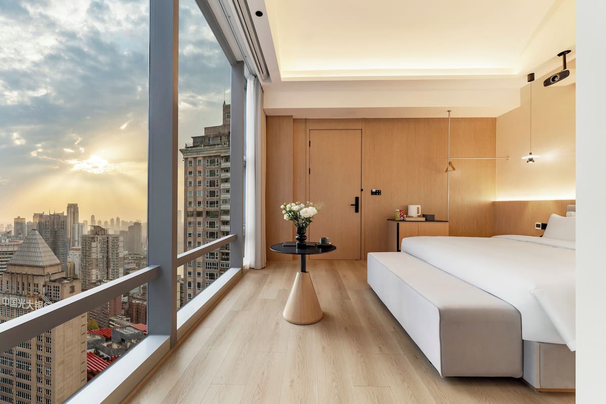 Ein modernes Hotelzimmer mit großem Fenster, einem Bett, einem Nachttisch mit Blumenvase und einer Holztür.
