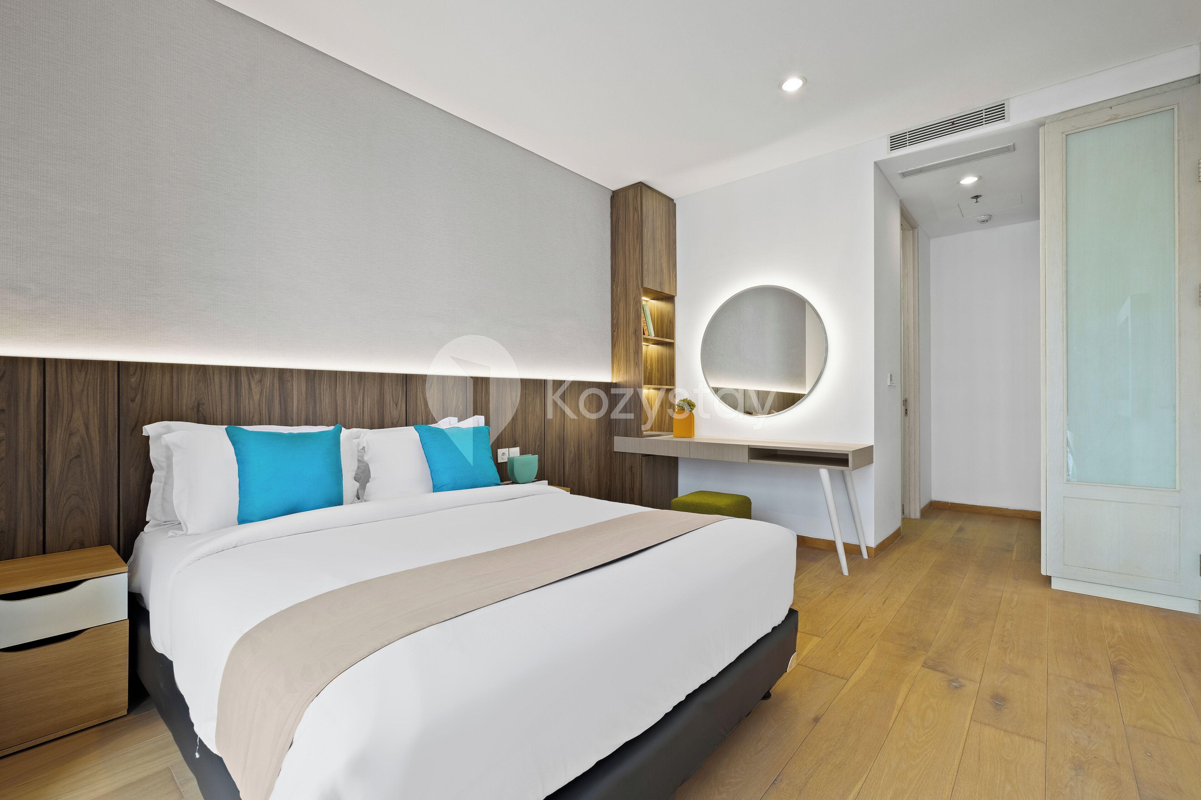 A modern hotel room with a large bed, a bedside table, a desk with a chair, a round mirror, and a wardrobe.