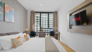 Une chambre d’hôtel moderne avec un grand lit, un bureau avec deux chaises, un téléviseur fixé au mur et une fenêtre avec des rideaux.