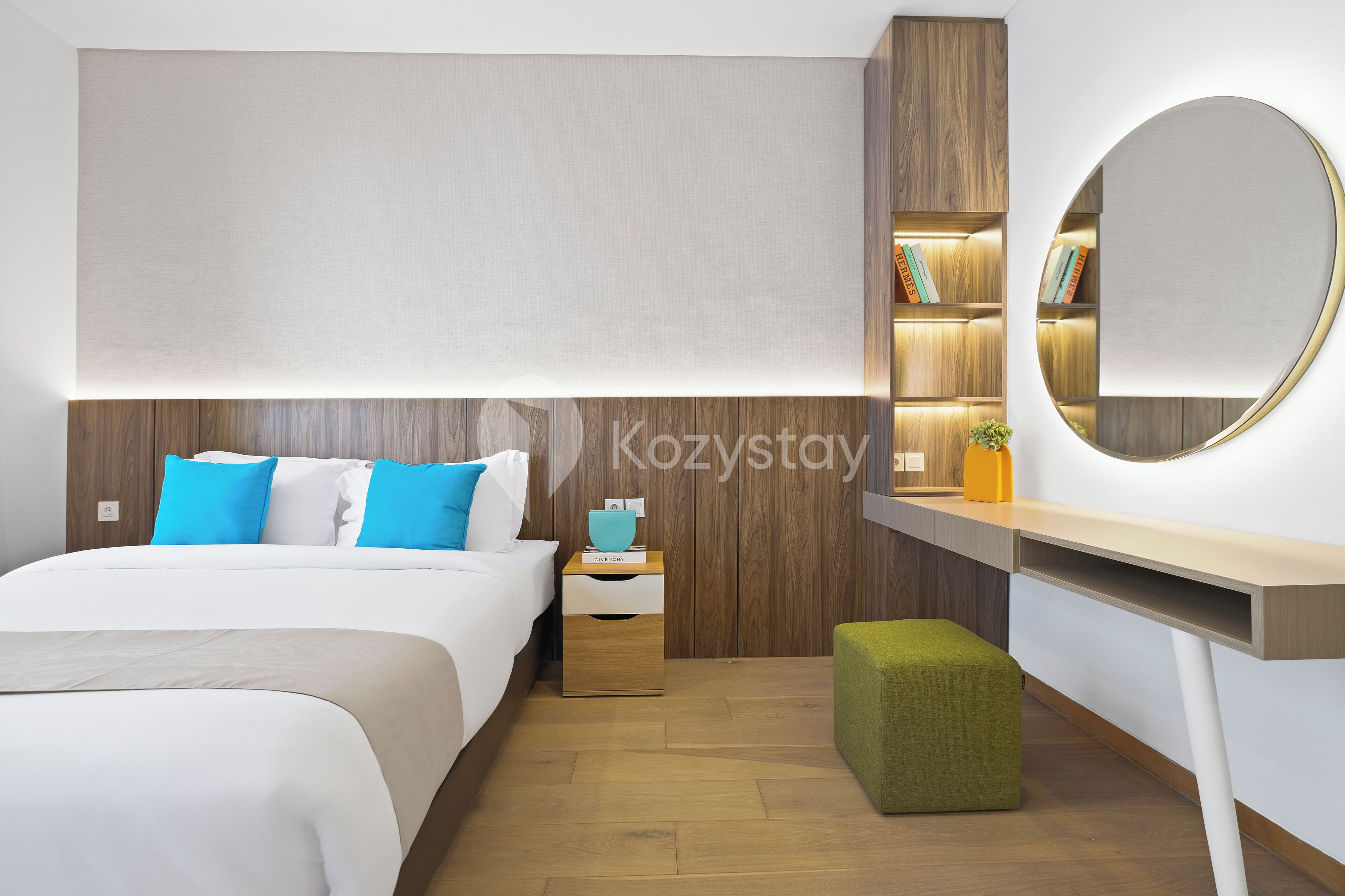 Een moderne hotelkamer met een groot bed, een nachtkastje, een groene poef, een houten hoofdbord, een wandspiegel en een inbouwboekenkast.