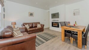A modern living room with a brown leather sofa, a wooden dining table with chairs, a flat-screen TV mounted above a fireplace, and a striped rug on the floor. - Luxury Apartment, City Break, 4B City Cottage Central Chester (Chester)