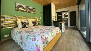 A modern bedroom with a wooden ceiling, a large bed with floral bedding, a cityscape painting, and a bathroom with a round mirror. - Lodge Vitré with Nordic bath for an unusual night in Dijon (DIJON)