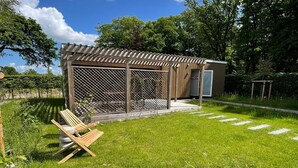 A small wooden structure with a covered patio, a fenced area, and a grassy lawn with a few chairs.