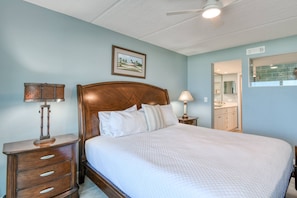 A bedroom with a wooden bed, two bedside tables, a ceiling fan, a framed picture on the wall, and a bathroom visible through an open door.