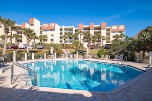 A swimming pool with a clear blue water, surrounded by a brick walkway. In the background, there are multi-story buildings with balconies and red-tiled roofs.