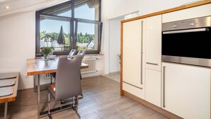 Une cuisine moderne dotée d’un four encastré et de placards, un coin repas avec une table et des chaises, et une grande fenêtre offrant une vue sur l’extérieur.