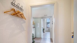 Un couloir avec un porte-manteau en bois, une porte blanche et une vue sur une pièce équipée d’un radiateur et d’une plante.
