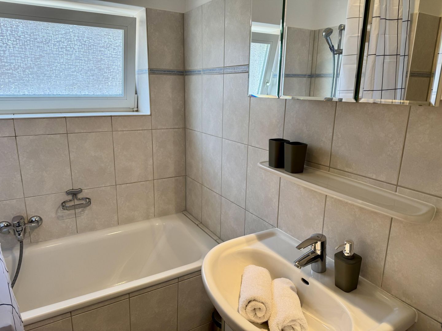A modern bathroom with a bathtub, a sink, and a mirror. There is a window above the bathtub, and a showerhead is visible in the mirror reflection.