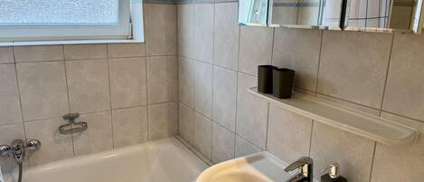 A modern bathroom with a bathtub, a sink, and a mirror. There is a window above the bathtub, and a showerhead is visible in the mirror reflection.