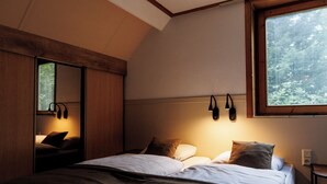 Une chambre à coucher avec un lit, des oreillers, une table de chevet, un miroir et une fenêtre donnant sur des arbres.