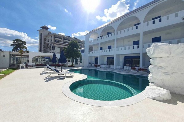 A swimming pool with a white rock feature, surrounded by a white building with multiple balconies and a clear sky.
