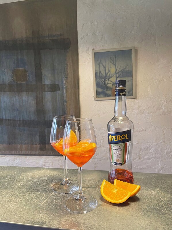 Una botella de Aperol con una rodaja de naranja sobre la encimera, dos vasos con rodajas de naranja y un cuadro en la pared.