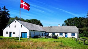 Un bâtiment blanc avec un toit de chaume, un mât sur lequel flotte un drapeau, et une table de pique-nique dans un champ herbeux.
