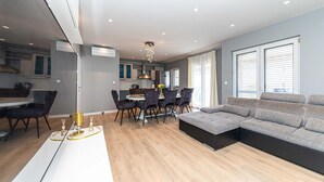 A modern living room with a sofa, dining table, and chairs. There is a kitchen area with cabinets and a window.