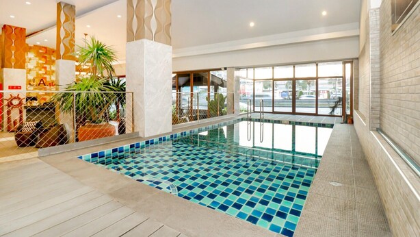 An indoor swimming pool with a tiled floor, surrounded by a wooden deck and a glass barrier. - Ultimate Twin Stay | Cozy, Smart Comfort (Pattaya)