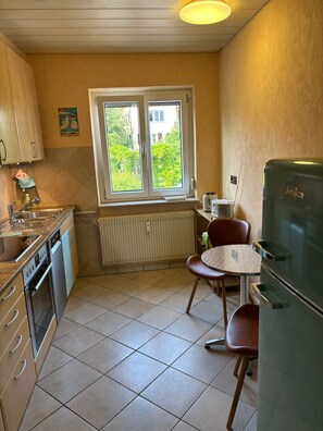 Une cuisine de taille réduite, dotée d’une fenêtre, d’un réfrigérateur, d’une table à manger avec des chaises et d’un radiateur.