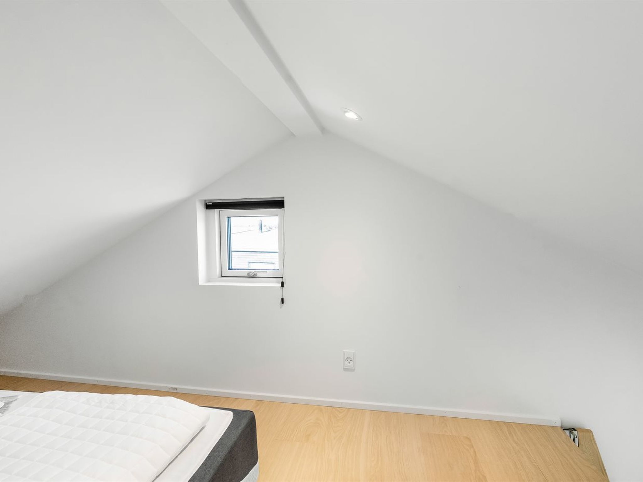 A bedroom with a sloped ceiling, a small window, and a bed with a white mattress.
