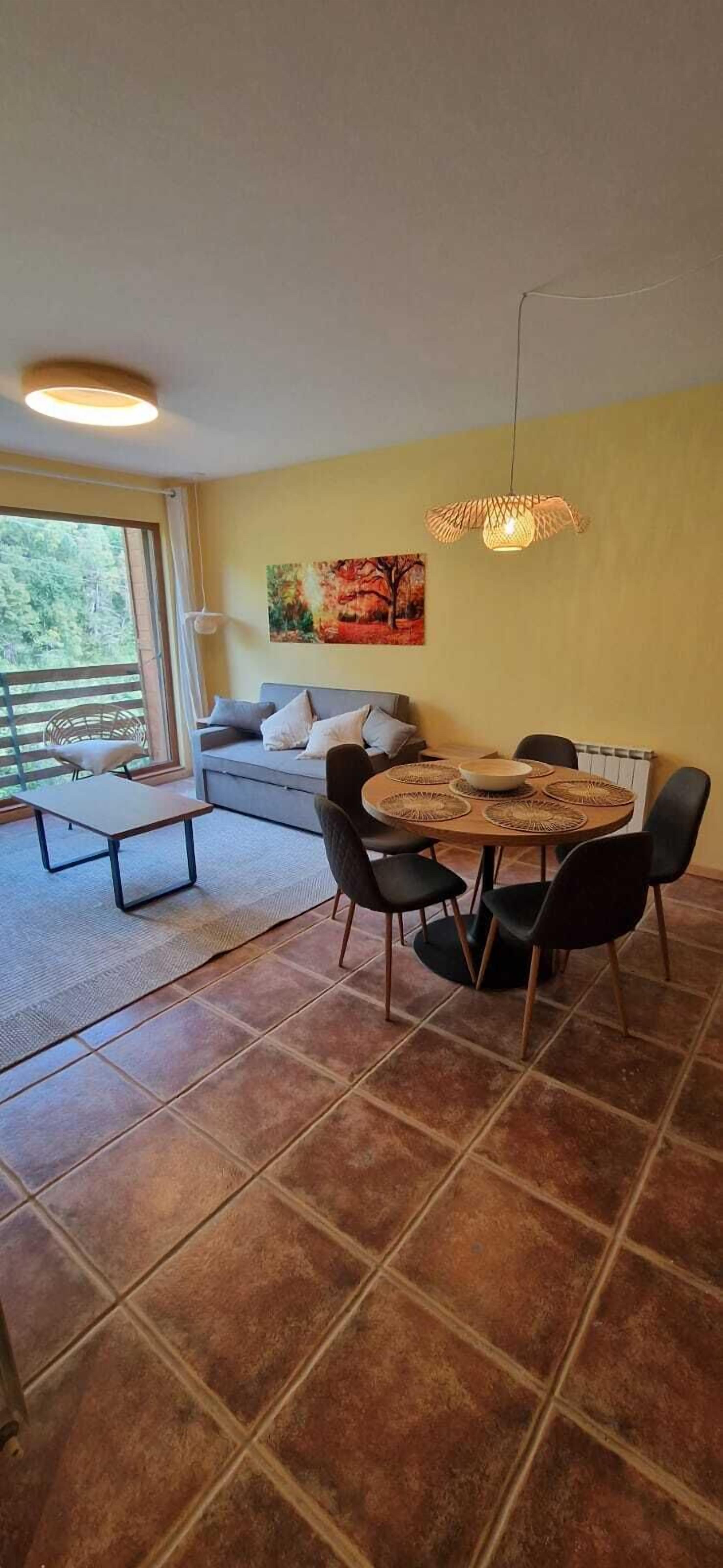 Una sala de estar moderna con un sofá, una mesa de comedor con sillas, una lámpara colgante y un ventanal con vistas a zonas verdes.