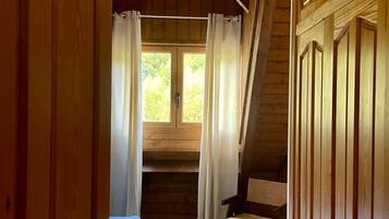 Interior de una cabaña de madera con dormitorio en altillo, un pequeño comedor y vistas al exterior con vegetación.