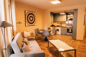 Ein modernes Wohnzimmer mit einer Couch, einem Esstisch und Stühlen. Im Hintergrund ist eine Küche mit Edelstahlgeräten zu sehen.