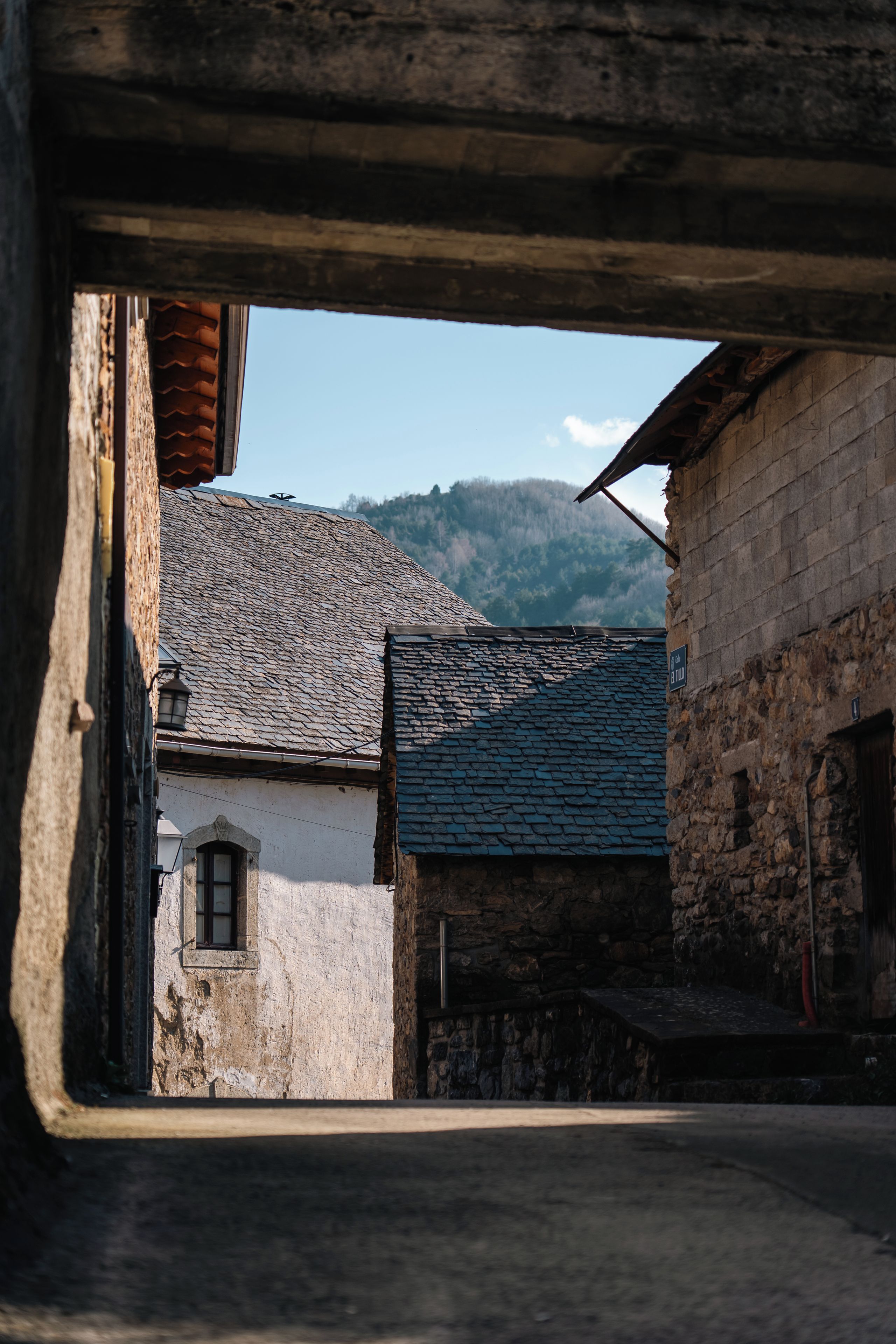 A narrow alleyway with stone buildings, one with a visible window and another with a sloped roof.