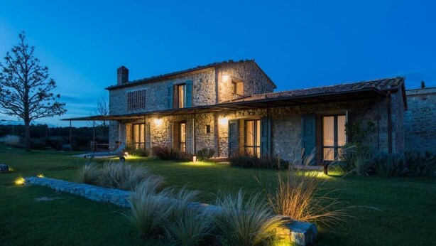 A stone house with a sloped roof, large windows, and a well-lit front yard with grass and plants. - Oak (Gavorrano)