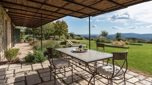 An outdoor patio with a table and chairs, a covered area, and a view of a grassy landscape with trees and hills. - Oak (Gavorrano)
