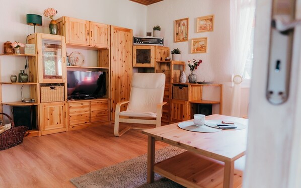 A cozy living room with wooden furniture, a television, a white armchair, and a dining table with a cup and a book.