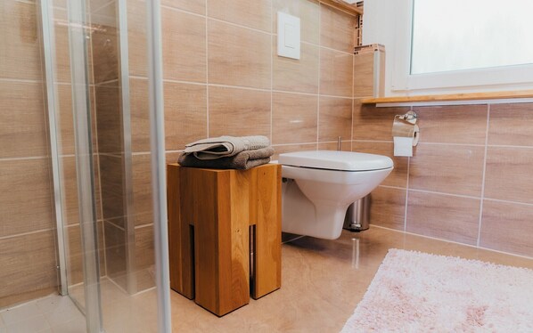 Un baño con inodoro, un taburete de madera con toallas, cabina de ducha de vidrio y una alfombra rosa.
