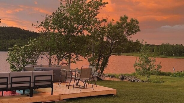 A deck with outdoor furniture overlooking a lake, surrounded by trees and a grassy area. - The Tidehouse, Relax with the whole family at this peaceful place to stay. (Laurenceton)