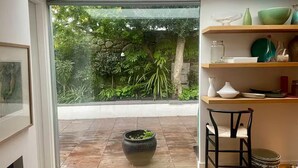 A room with a wooden floor, a black chair, a large potted plant, and a view of a garden through a glass door.