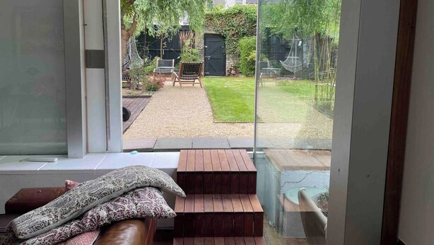 A modern room with a large sliding glass door leading to a garden. Inside, there is a wooden bench and a cushioned seating area.