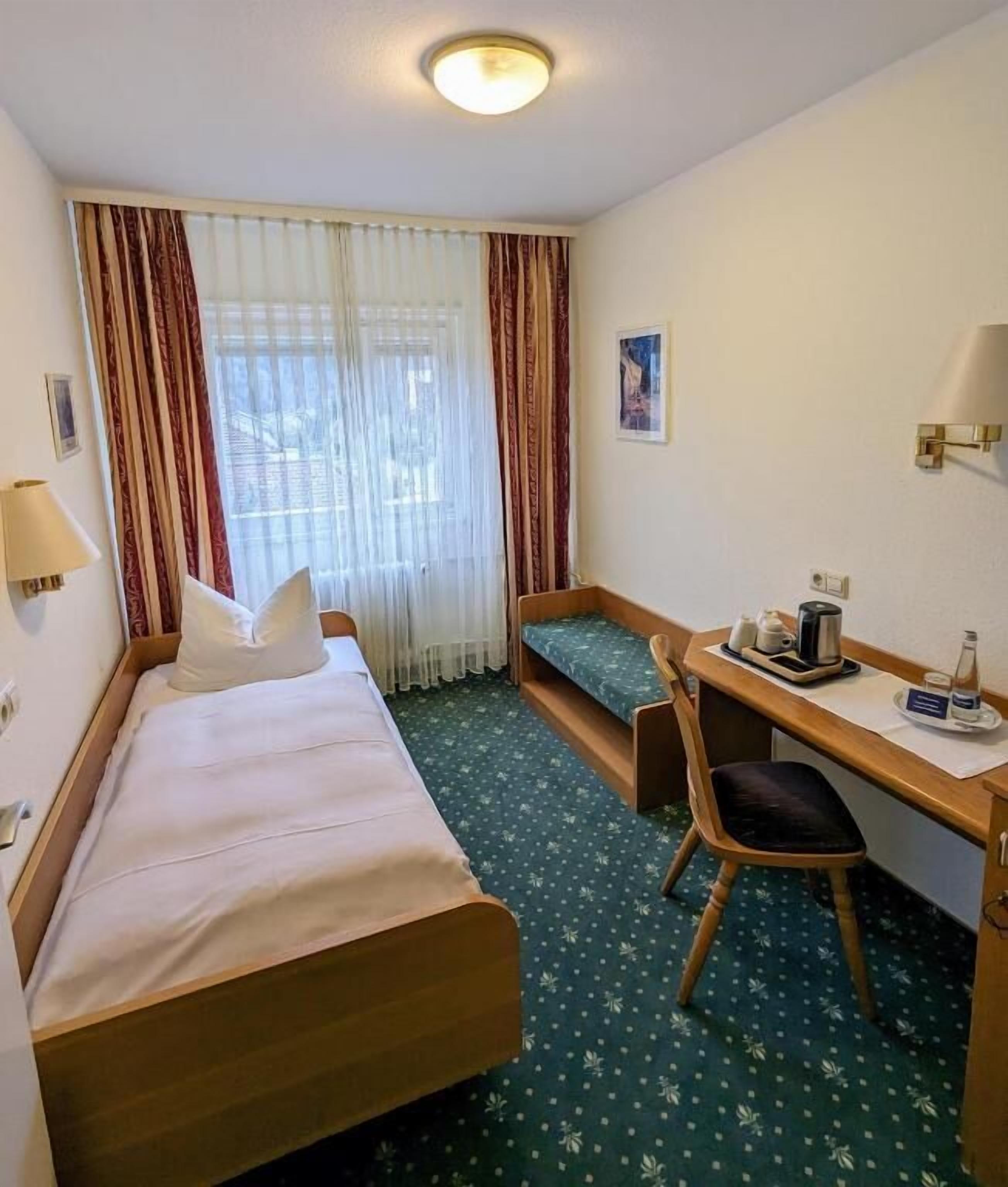 A hotel room with a bed, a desk, a chair, a window with curtains, a lamp, and a small table with a kettle and cups.