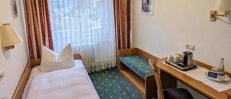 A hotel room with a bed, a desk, a chair, a window with curtains, a lamp, and a small table with a kettle and cups.