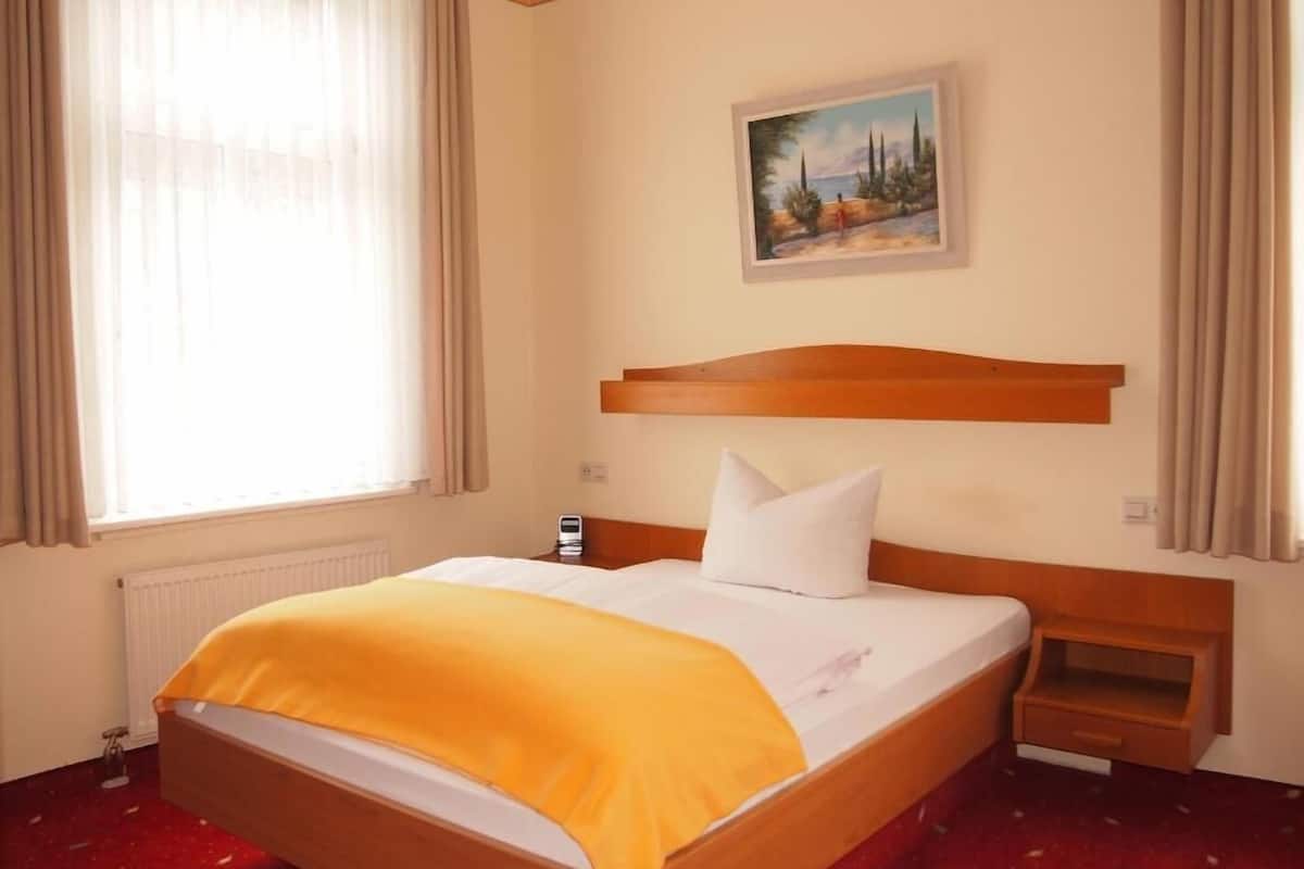 A hotel room with a wooden bed, a painting on the wall, and a window with curtains.