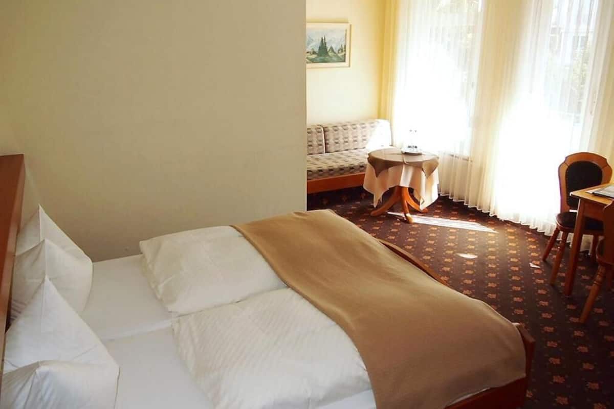 A hotel room with a bed, a small table, a chair, a sofa, and a window with curtains.
