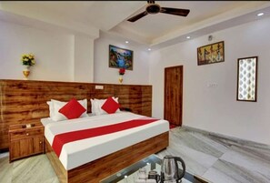 A hotel room with a wooden headboard, a bed with white bedding and red accents, a ceiling fan, and artwork on the walls. - Hotel The Grand (New Delhi)
