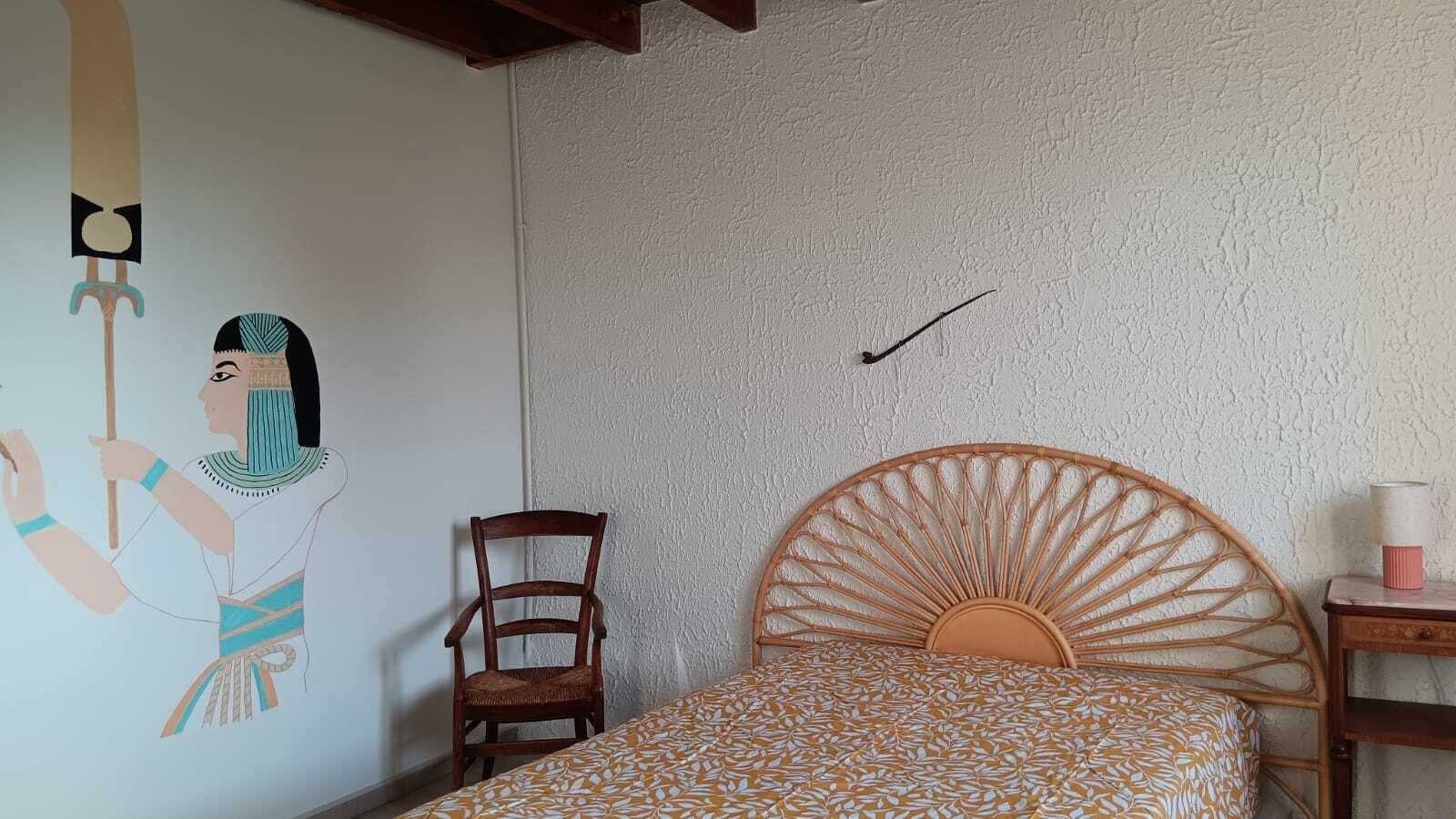Een slaapkamer met een bed, een stoel, een muur met een decoratief schilderij en een klein tafeltje met een lamp.